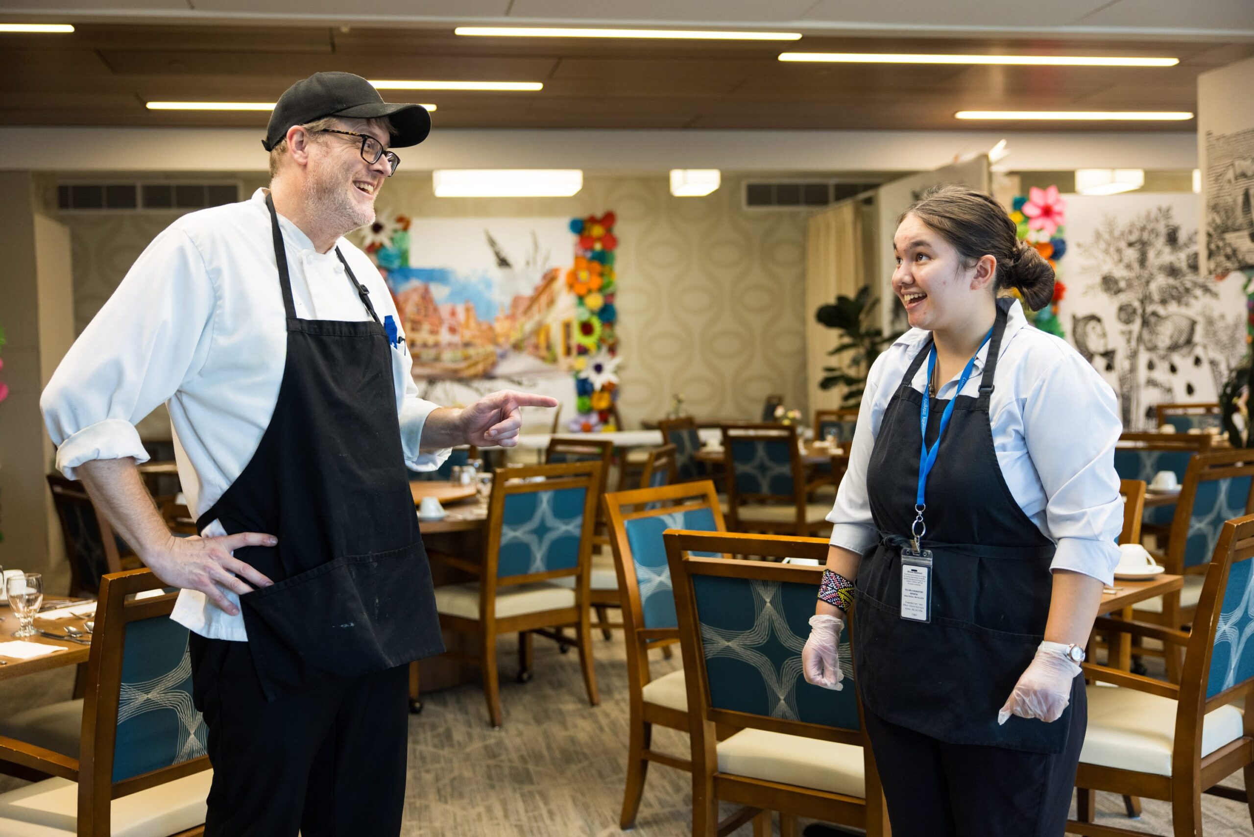 Two food service coworkers wearing black aprons talking and smiling at each other.